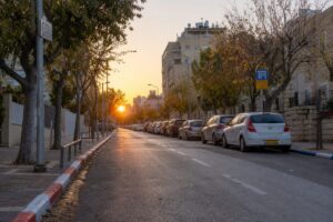 A realistic photograph of an urban street in an Israeli city during the early hours of the morning, just as the sun begins to rise. The street is lined with parked cars, positioned between distinctively painted curbs: a mix of blue-and-white, red-and-white, and plain sections. The lighting casts a warm glow over the scene, highlighting the freshly painted curb colors and the quiet stillness of the city before it wakes up. Trees with early autumn leaves and streetlights add depth to the scene. High definition, capturing intricate details like the texture of the curb and the reflections on the car windows.