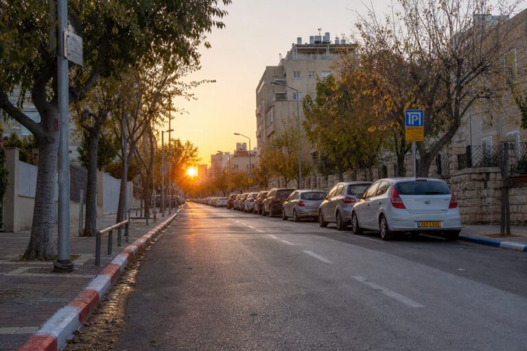 A realistic photograph of an urban street in an Israeli city during the early hours of the morning, just as the sun begins to rise. The street is lined with parked cars, positioned between distinctively painted curbs: a mix of blue-and-white, red-and-white, and plain sections. The lighting casts a warm glow over the scene, highlighting the freshly painted curb colors and the quiet stillness of the city before it wakes up. Trees with early autumn leaves and streetlights add depth to the scene. High definition, capturing intricate details like the texture of the curb and the reflections on the car windows.