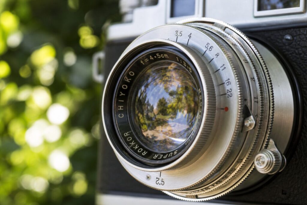 A realistic photograph capturing a close-up of a colorful vintage camera lens set against a soft-focus backdrop of lush greenery. The lens reflects an outdoor scene, bathed in natural sunlight, with intricate details visible in the lens' metal and glass surfaces. The depth of field is shallow, emphasizing the lens while softly blurring the background. The image should be in high definition, capturing the textures and reflections on the lens with precision.