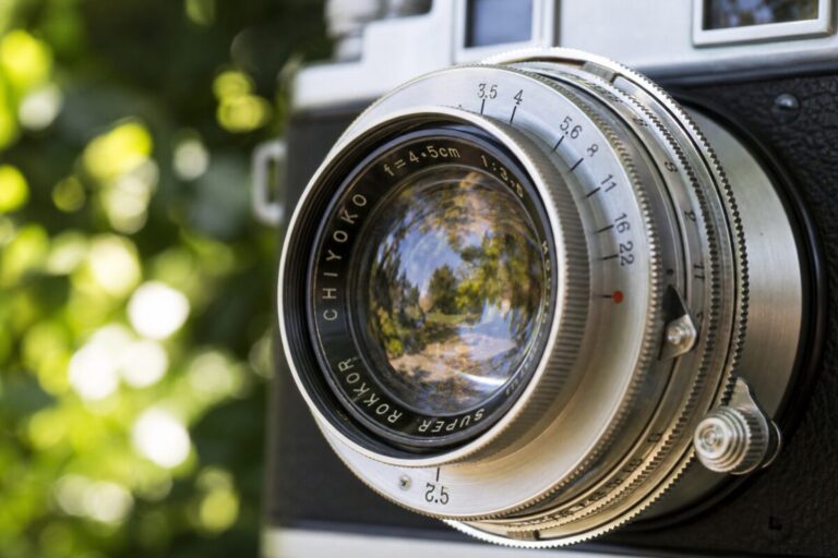 A realistic photograph capturing a close-up of a colorful vintage camera lens set against a soft-focus backdrop of lush greenery. The lens reflects an outdoor scene, bathed in natural sunlight, with intricate details visible in the lens' metal and glass surfaces. The depth of field is shallow, emphasizing the lens while softly blurring the background. The image should be in high definition, capturing the textures and reflections on the lens with precision.